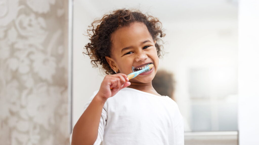 A portrait photo of a young child with curly brown hair, wearing a white t-shirt, happily brushing her teeth in a brightly lit bathroom.
